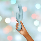 A hand with painted nails holds the Femme Funn Balai Rabbit Vibrator - Light Blue by Femme Funn, showcasing its blue dual-stimulation design with a curved main shaft and smaller external stimulator. The background is blurred with bokeh lights in various pastel colors, adding to the festive and dreamy appearance.